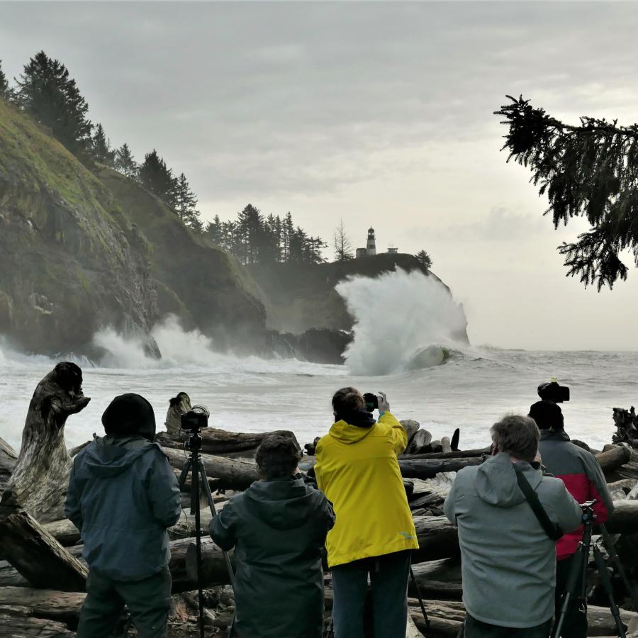 Eight people stand on a rugged shoreline holding cameras and photographing the surf crashing high against the cliffs at Cape Disappointment State Park. North Head Lighthouse can be seen in the distance on top of the cliff where the tall white wave is crashing hardest.