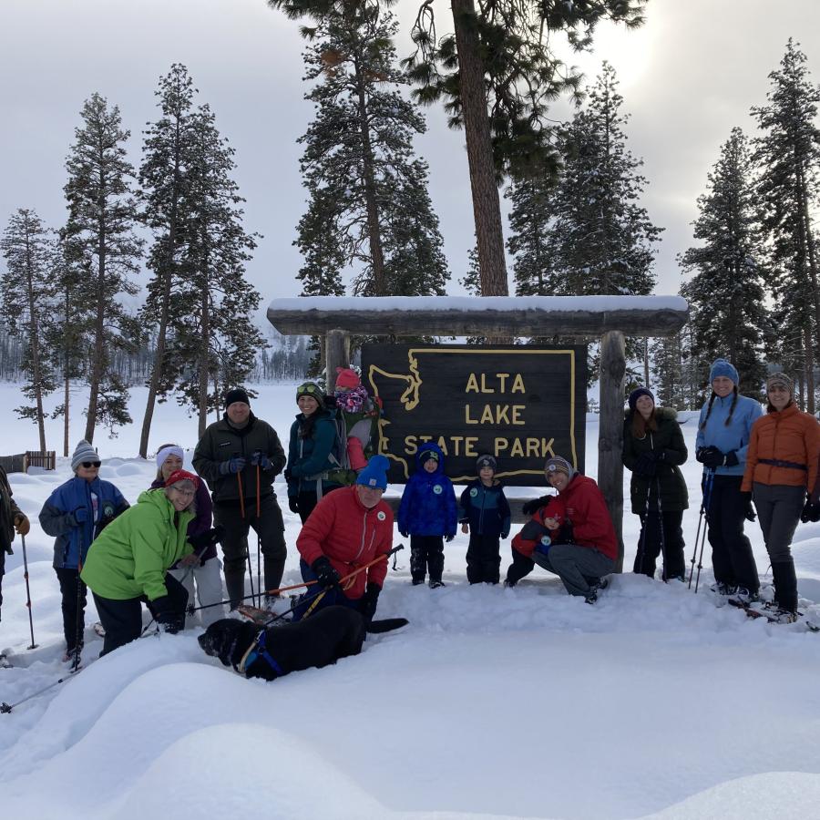 A group of people in winter clothing and gear stand in the snow next to a wood sign reading Alta Lake State Park