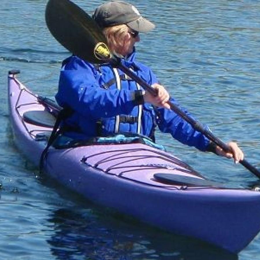 A kayaker in a blue jacket with a black hat sits in a purple kayak, using black paddles to move.