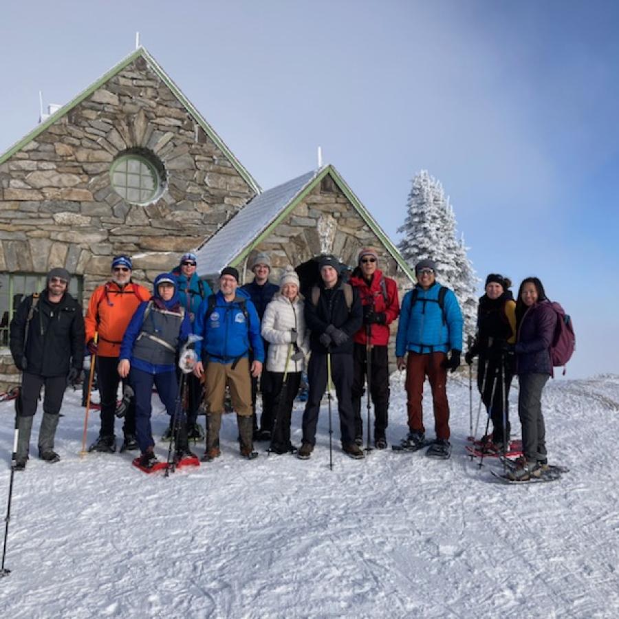 A group of snowshoers in colorful winter clothes stands on a field of fluffy snow in front of a snow covered stone cabin. Pale clouds rise into a clear blue sky behind them.