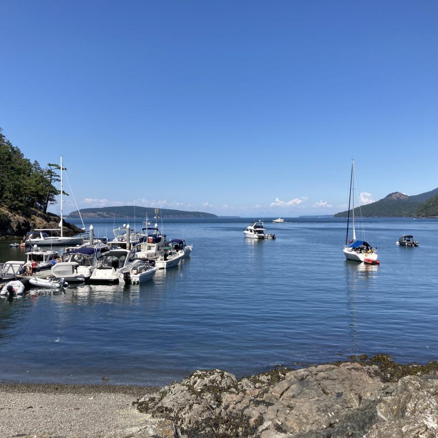 Course, grey sand beach with grey rocks on the right meet the blue water in the foreground. On the left are numerous motor boats tied to a long dock, the edge of a treed, rocky hillside sit behind them. Several motor and sail boats sit in the bay looking towards the open water. A hand full of treed islands in front of cloudy horizon with a blue sky in the background.