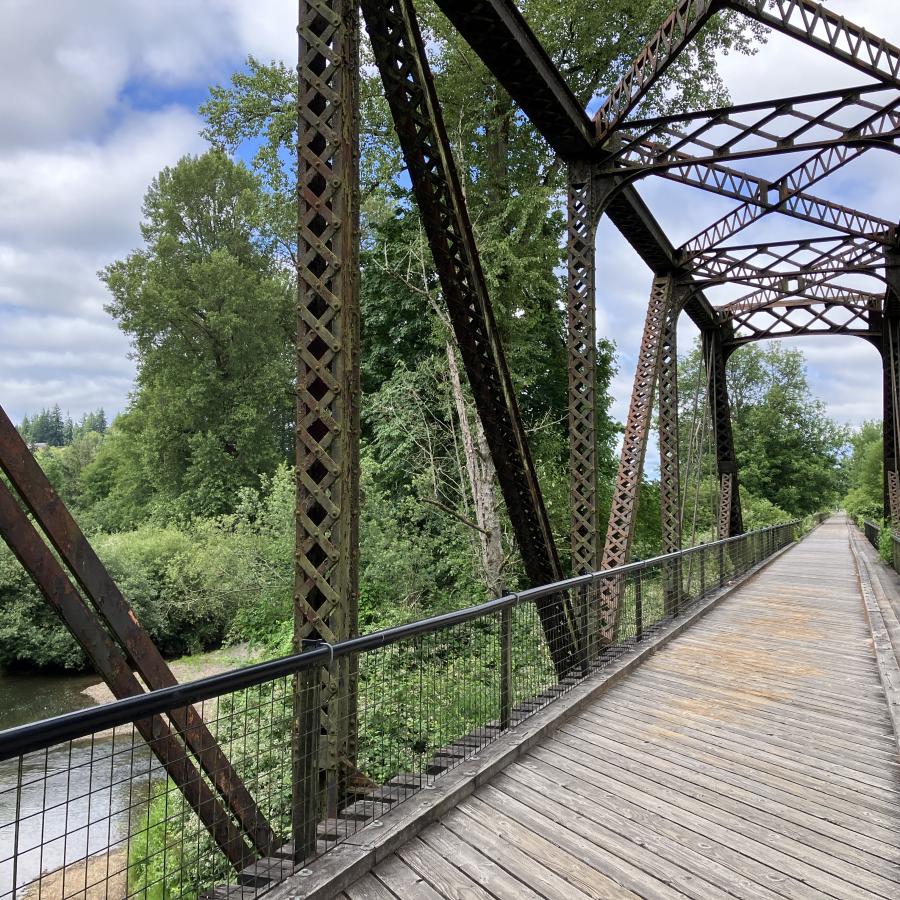 Looking down an old iron train trestle bridge over a river that has been converted to a bike path. It has a wooden deck and is surrounded by green trees and is under a blue sky.