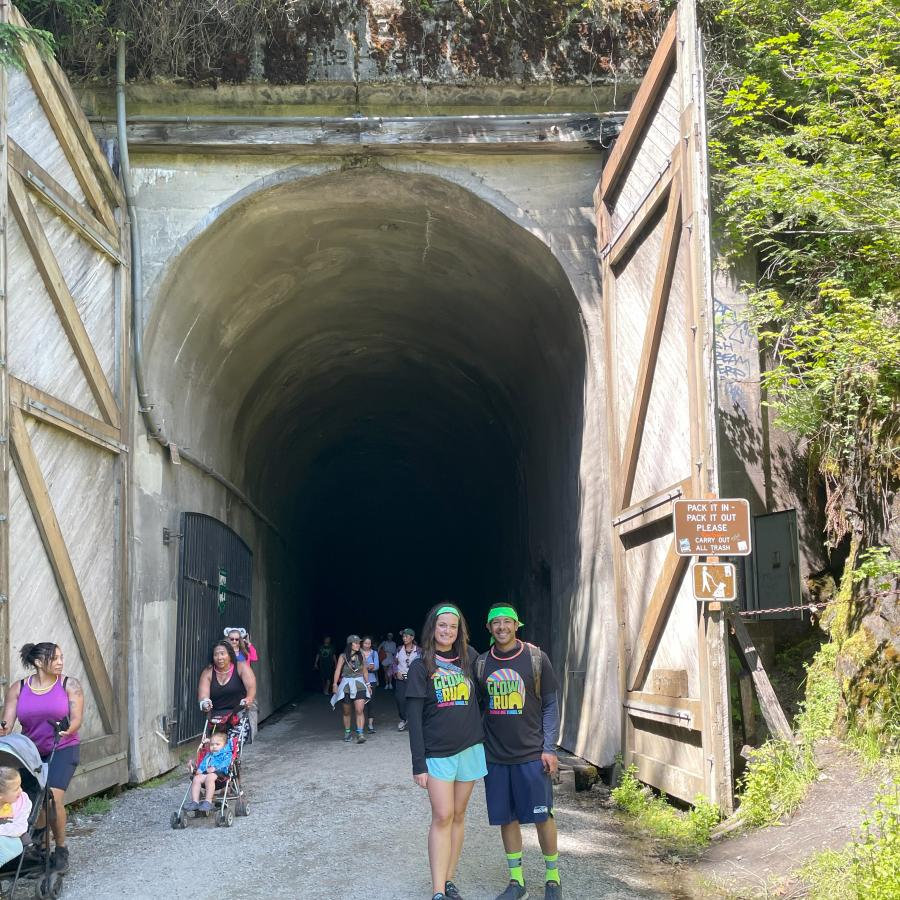 A couple smiles in front of a tunnel entrance where several other people are walking in and out
