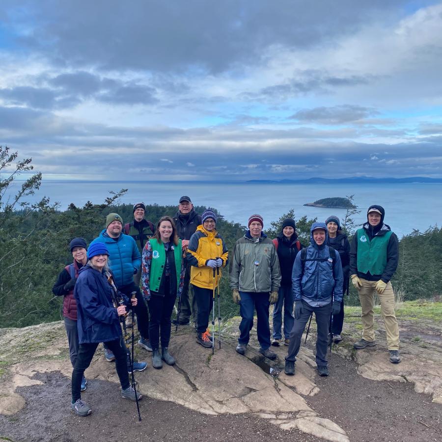 Group of hikers standing on a summit with evergreen trees and water in background
