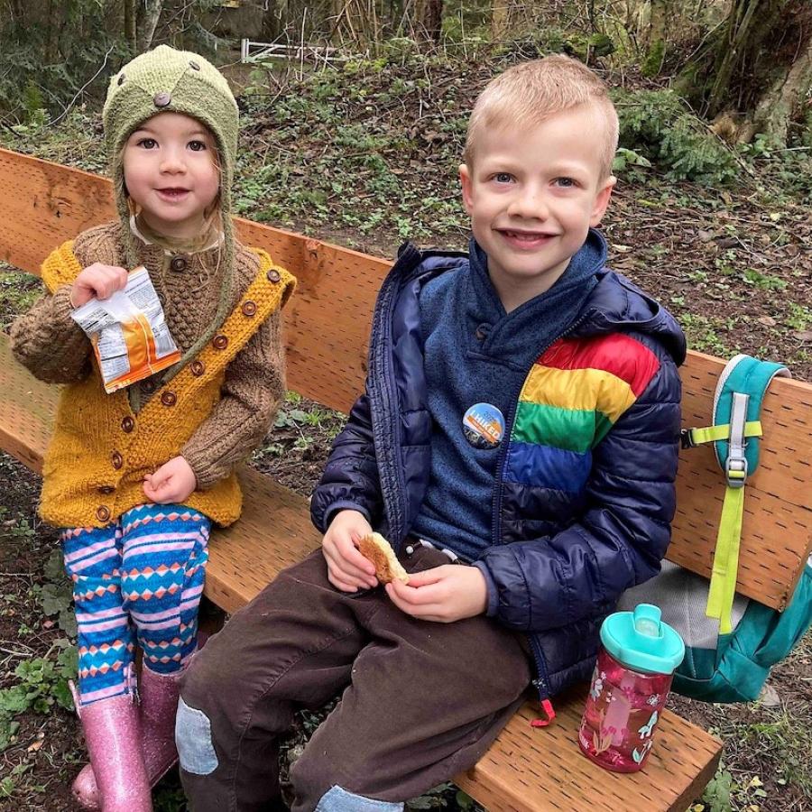Young girl and boy bundled in warm jackets with First Day Hike buttons sitting on park bench eating snacks