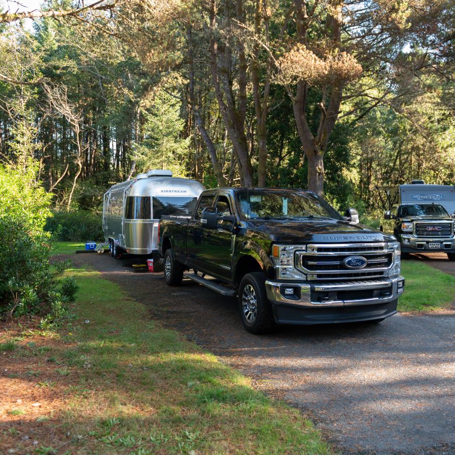 A truck and camper sit on a concrete driveway at the campsite surrounded by trees, bushes and some grass. 