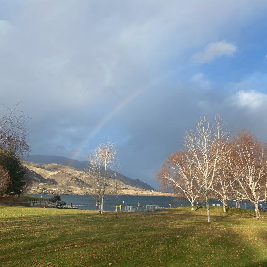 This is an image of a large grassy field leading to the water with a mountain landscape in the background. The leaves on the trees are changing to golden orange. A rainbow glows from the far mountain scape over the water. 