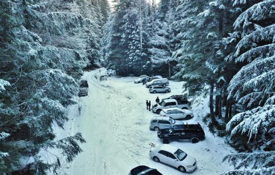A forest road leading to a parking area that comfortably fits 12 vehicles , both covered in snow, surrounded by snow-dusted trees on both sides. 