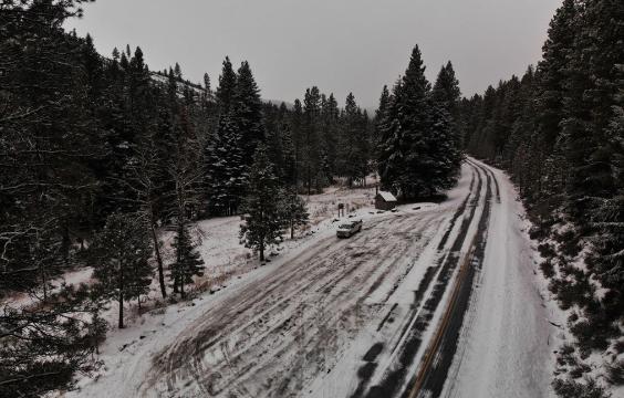 A plowed parking pull-off area next to a plowed highway flanked by snow-dusted pine trees.