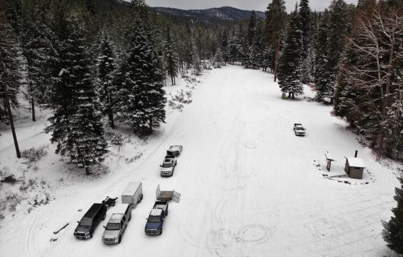 Several trucks parked on a long snow-covered parking area surrouned by pine trees. A pit toilet hut sits in the lower right corner.