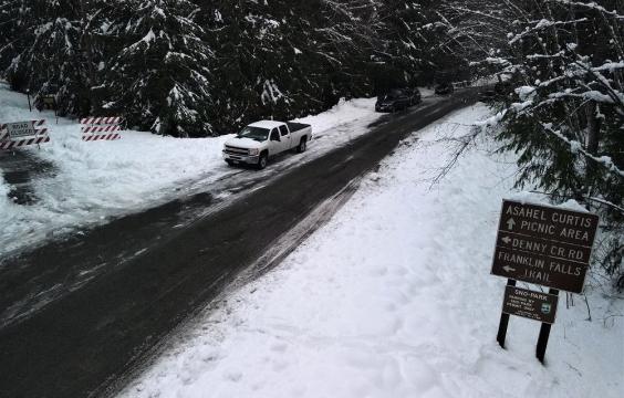 several cars and a truck parked on the side of a plowed access road with snow on both sides and a sign pointing to picnic area