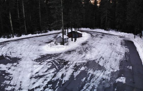 Oval paved parking lot covered thinly with snow and ice bordered by piled up snow and thick pine trees. A warming hut and pit toilet hut stand in the middle.