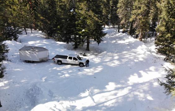 A snowy forest road with a pull-out surrounded by pine trees. A truck with a trailer stands in the pull-out.