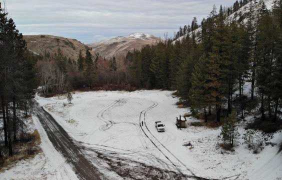 An open snow-covered parking area lined with trees next to a plowed highway with a pit toilet hut on the edge. Hills lightly dusted with snow lie in the background.