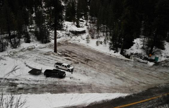 Plowed parking area with two trucks in the middle, next to a plowed highway surrounded by pine trees. A porta-potty stands in the corner.