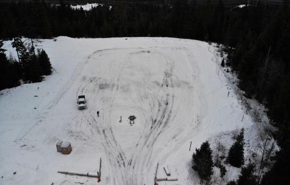 A rectangular parking area covered with a layer of thin snow and tire tracks. Two porta-potties sit in the lower left corner.