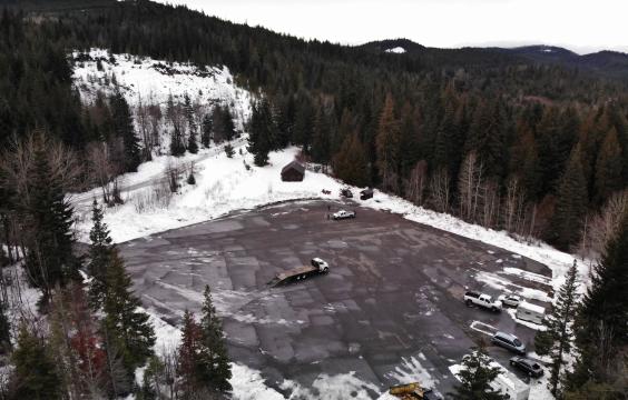 Large square paved parking lot plowed clean surrouned by pine trees next to a forest road with light snow. A warming hut and pit toilet hut stand in the upper right corner and several trucks are parked along the right edge of the lot.