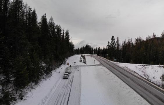 A plowed highway turns onto a plowed forest road covered by a thin layer of snow. A truck with a trailer and a porta-potty stand next to an information board.
