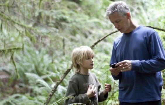 A child and an adult stand in a mossy forest. They are looking at their GPS units.