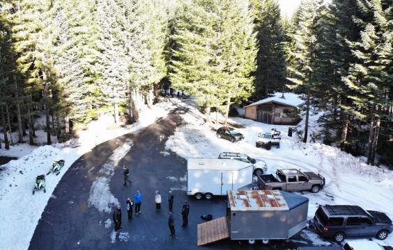 A snow covered forest road opens onto  a wider plowed parking area surrouned by pine trees. A warming hut and several trucks with trailers lie on one side of the lot and two snowmobiles lie on the other side.