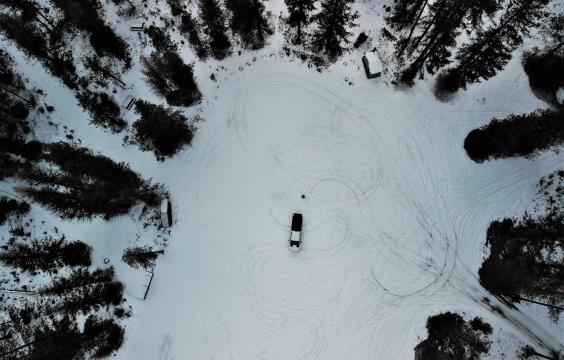 Overhead view of an open snow-covered parking area surrounded by pine trees.A pit toilet hut and picnic tables sit along the edge.