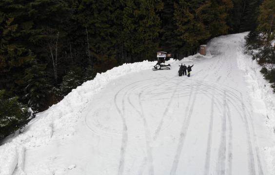 Snow-covered forest road opening onto a parking area covered by snow and tire tracks. A porta-potty and two snowmobiles and their riders are on the left edge of the lot.