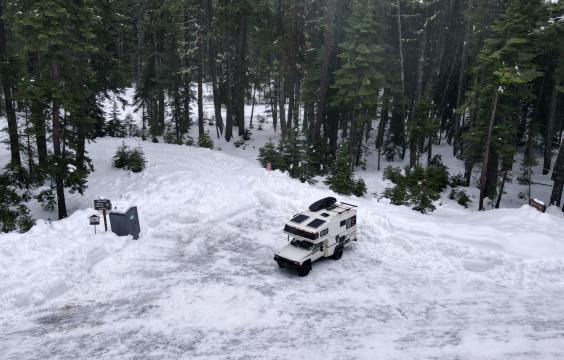 Parking area covered by bumpy ice and snow surrouned by thick snow-covered ground and pine trees. A truck is parked in the middle and a porta-potty sits on the left edge of the lot.