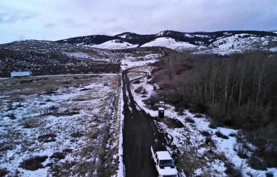 A plowed highway runs between a forest and a meadow with patchy snow with snowy hills in the background.