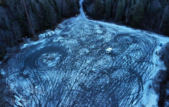 A large parking area with thin layer of ice and snow overrun by numerous tire tracks, surrounded by a thick dense forrest. A porta-potty sits in the corner.