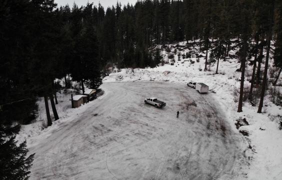 A parking lot covered by ice bordered by pine trees with a pit toilet hut on the left side and a truck with a trailer on the edge.