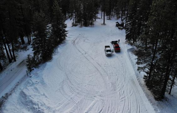 Overhead view of a snow-covered parking area lined with pine trees next to a plowed highway. A pit toilet hut sits in the upper right corner.