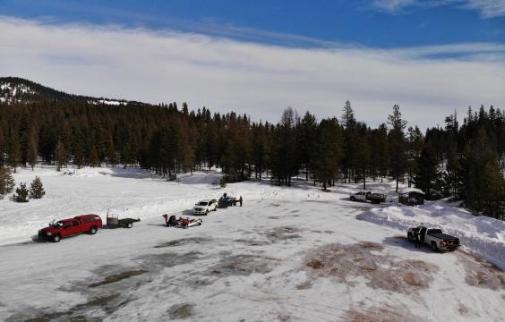 Large plowed parking lot with vehicles and snowmobiles and a pit toilet hut in the corner.