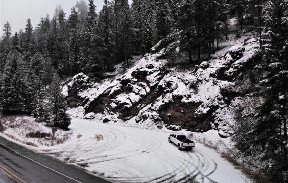 A snow-covered turn-off from a plowed highway with a wall of sloping rocks and trees on the right side dusted in snow.