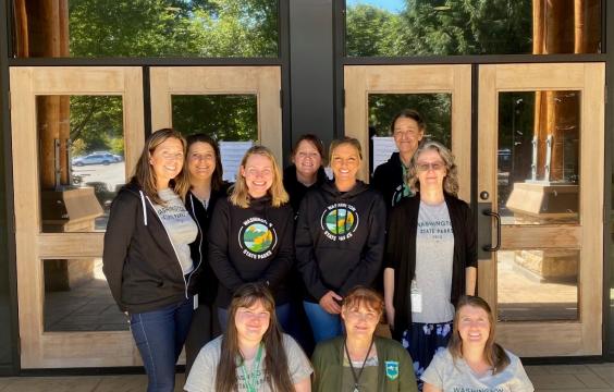 Seven members of the State Parks Human Resources team stand in front of the agency headquarters doors. Three more team members kneel in front.
