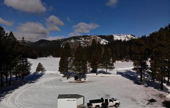 A truck with a trailer sits on the edge of an oval snow-covered parking area surrounded by evergreen trees with snowy hills in the background.