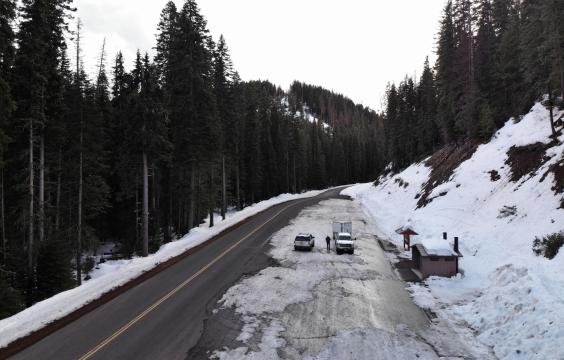 A ice-covered pull-off parking area next to a plowed highway lined with evergreen trees. A pit toilet hut stands on the side of the lot.
