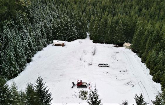 Overhead view of a square parking area covered by thick snow and bordered by a thick pine tree forest. A warming hut sits in the upper left corner and pit toilet hut sits on the upper right corner.