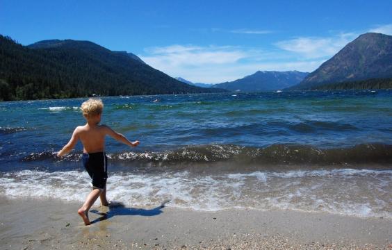 A child runs on a sandy beach toward the lacy white foam of the receding surf at Lake Wenatchee State Park. The water is a deep blue, reflecting the clear sky. In the distance are the dark slopes of the foothills.
