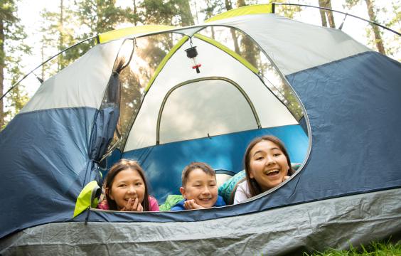 Three children poke their heads out of a tent. They are smiling.