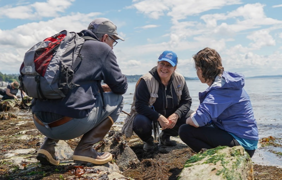 Three people squatting on the beach engaged in a discussion. In the middle is the Seattle Aquarium Beach Naturalist who is a feminine appearing person in a blue Seattle aquarium hat wearing a khaki colored vest and speaking to a masculine looking person to their left in a grey sweatshirt and jeans with a red and grey backpack. To the right of the beach naturalist is a feminine appearing person with brown hair and a purple jacket with black plants.