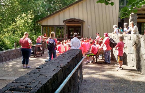 A large group of children sit outside a building and listen while a park ranger speaks.